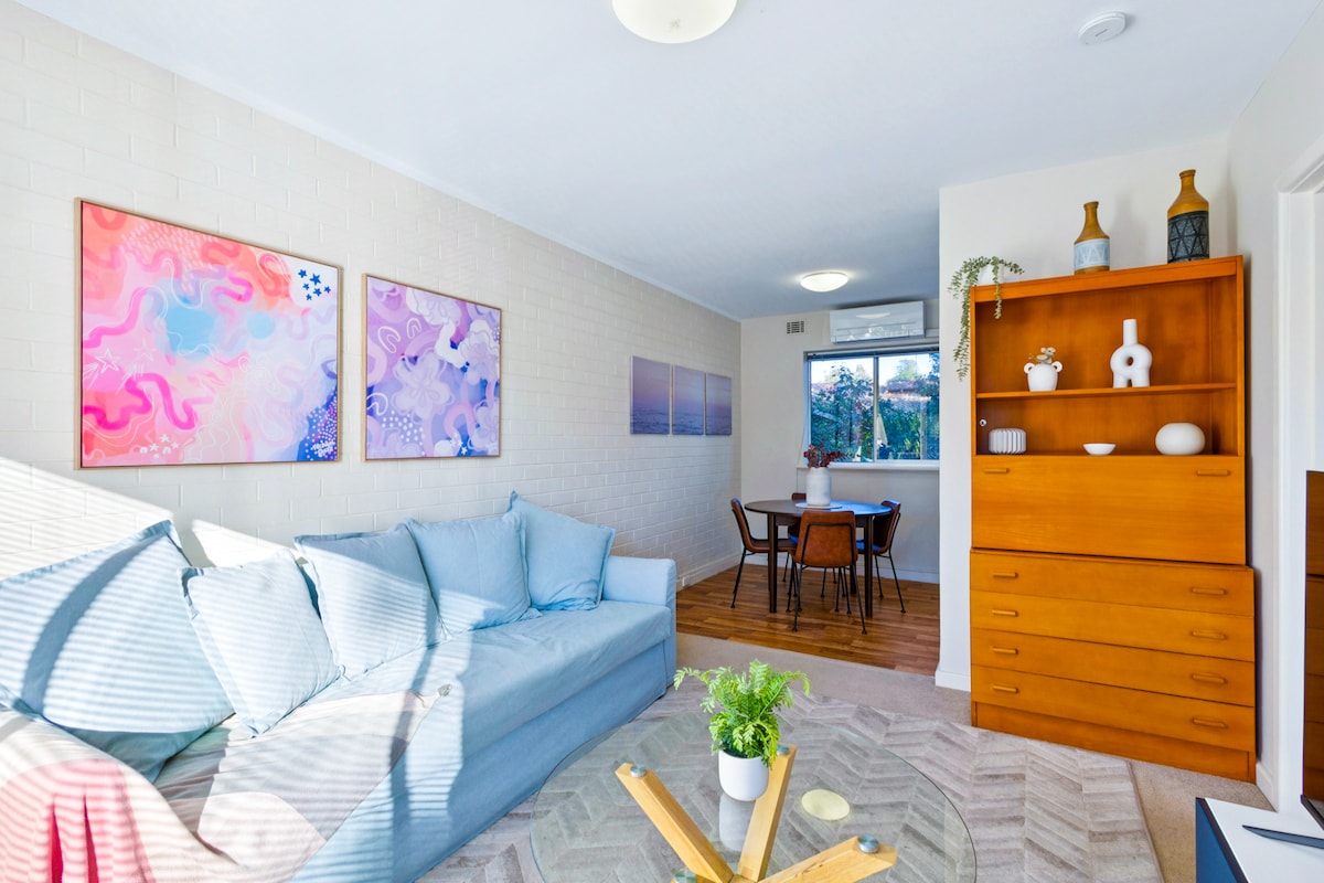 A comfortable living area is depicted with a light blue sofa that complements the wooden furnishings. A coffee table sits center stage, while two colorful wall art pieces add interest. A dining area for four is visible in the background, bathed in natural light.