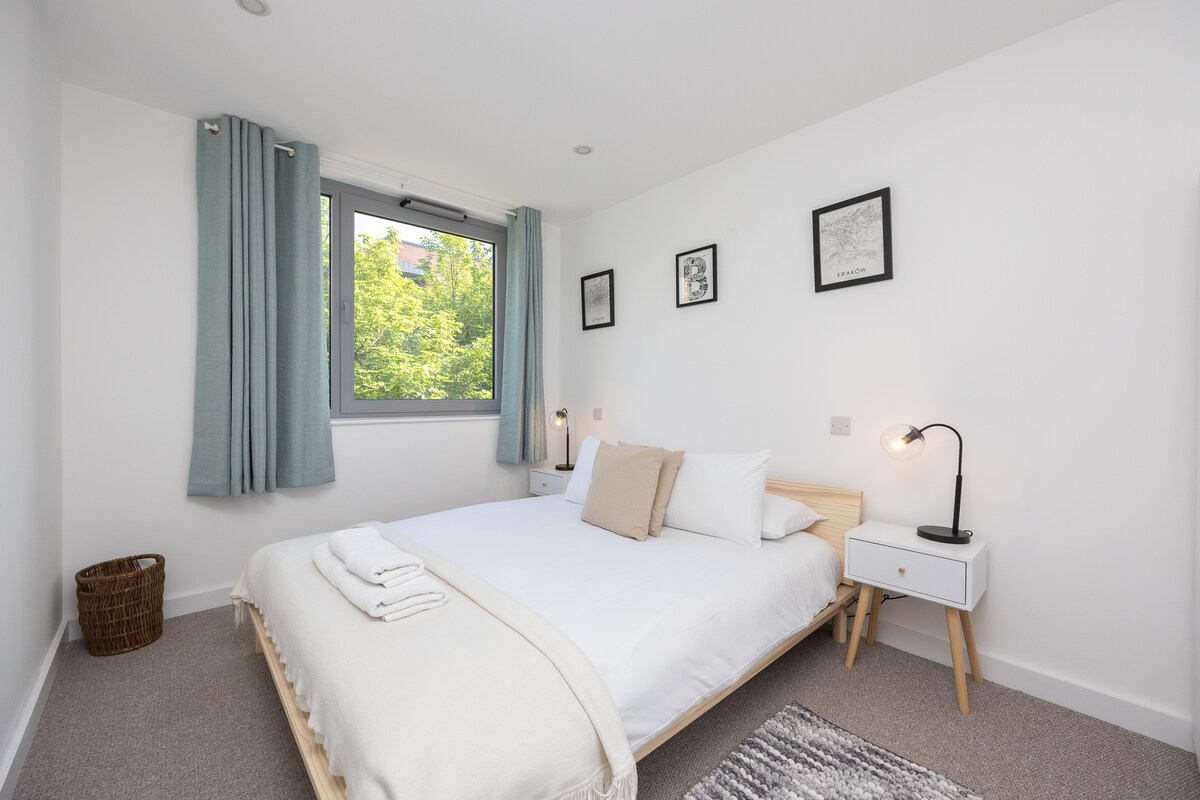 A bright bedroom features a spacious bed with crisp white linens and soft neutral accents. Large windows allow natural light to enter, with views of greenery outside. A bedside table with a lamp complements the minimalistic decor, while a basket adds a touch of warmth.