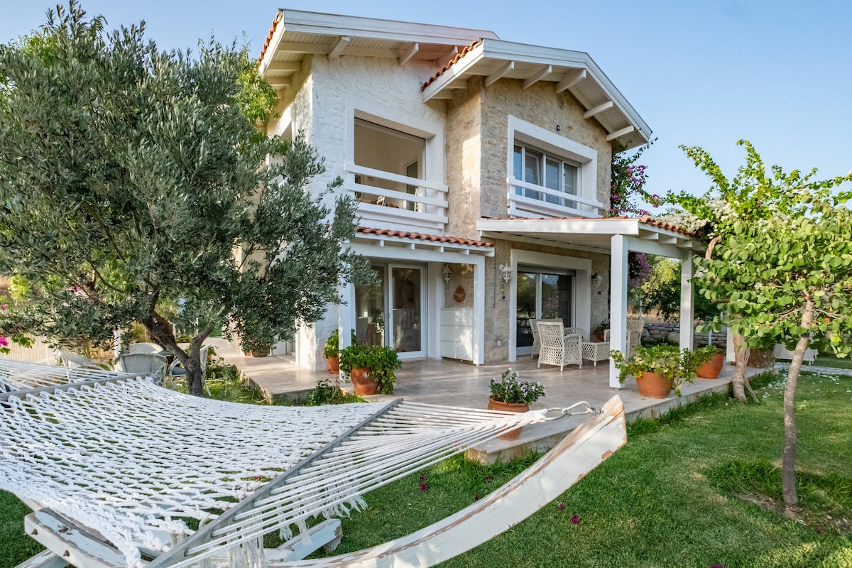 The two-story house is surrounded by greenery, complemented by a charming porch with seating. A hammock is visible in the foreground, inviting relaxation. Large windows enhance natural light, highlighting the earthy tones of the exterior and the well-kept garden.