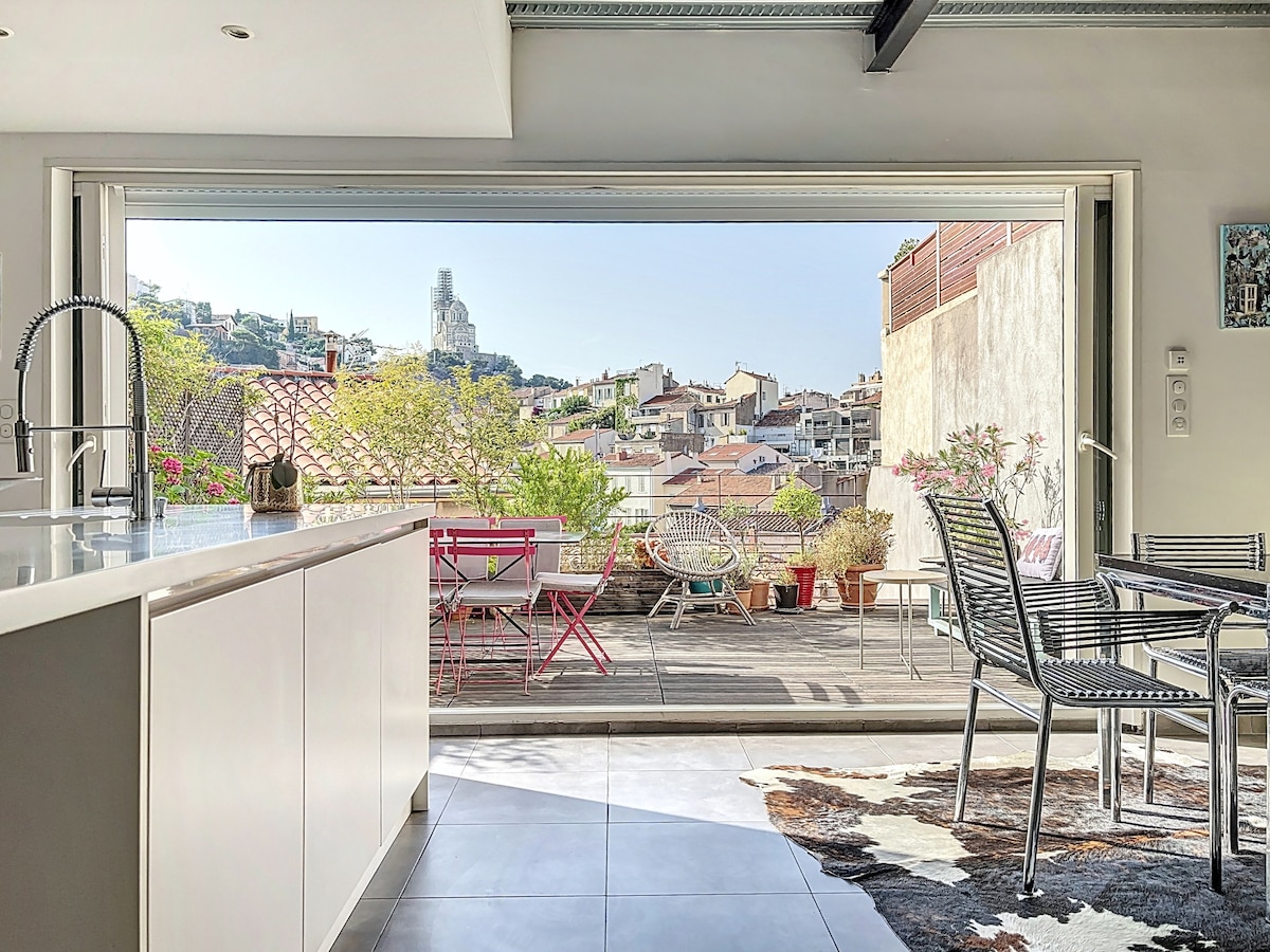 A modern kitchen opens to a spacious terrace with a view of the city. Light wooden cabinetry complements the streamlined design. An arrangement of colorful outdoor furniture, including metal chairs and a table, enhances the inviting outdoor space. The iconic Notre-Dame de la Garde is visible in the background.
