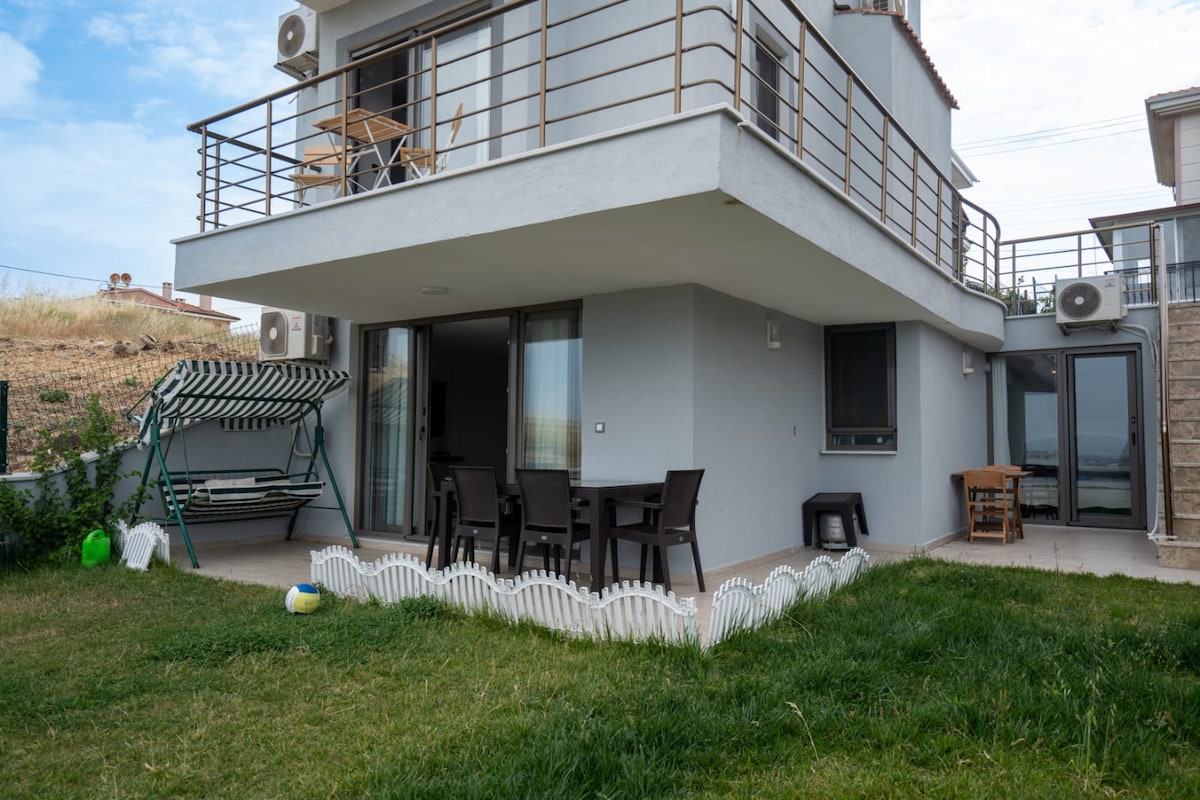 A private garden area features a swing and outdoor seating with a table and chairs. The exterior of the villa includes large glass doors for natural light. The grass is well-maintained, and a white picket fence outlines the garden space.