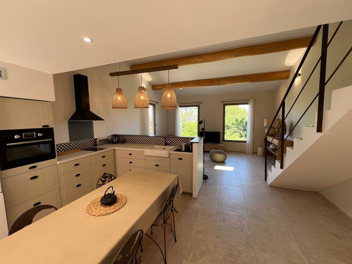 The kitchen and dining area features a spacious layout with light-colored cabinets and modern appliances. Natural light streams through the window, illuminating the area, while wooden beams add a touch of warmth. A dining table with tasteful seating is positioned near the staircase.