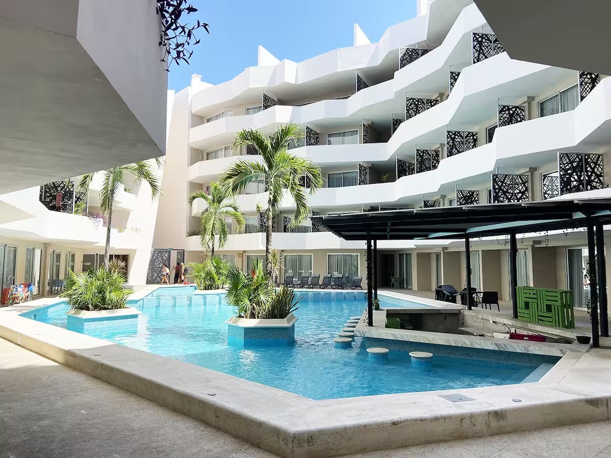 A tranquil outdoor area is displayed, featuring a curvilinear pool surrounded by lush palm trees and tropical plants. The modern building showcases a series of balconies and large windows, creating an open atmosphere. Lounge chairs are positioned near the pool for relaxation.