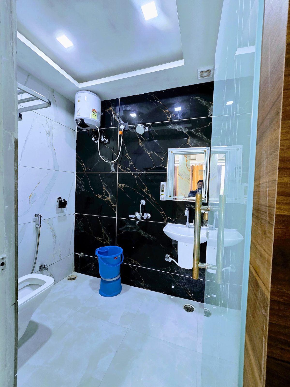 A modern bathroom features black marble walls, accented by contrasting white tiles. A shower area is visible along with a toilet and a washbasin, while a blue bucket adds a pop of color to the space. Bright overhead lighting provides a well-lit atmosphere.