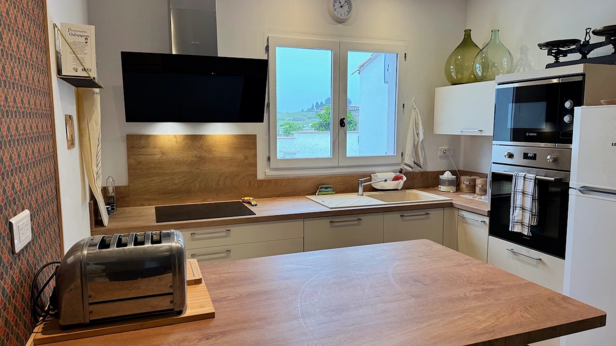 A modern kitchen with wooden cabinetry and a spacious countertop is displayed. A toaster, stovetop, and wall-mounted oven are visible, along with a large window that allows natural light to illuminate the area. Green glass vases and kitchen essentials are neatly arranged on the shelves.
