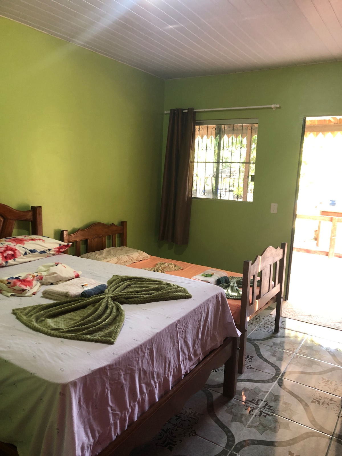 A brightly painted green room features two beds, one with decorative linens and a towel arranged in a bow shape. Natural light enters through a window, illuminating the space. A door leads to an outdoor terrace, enhancing the room's airiness.