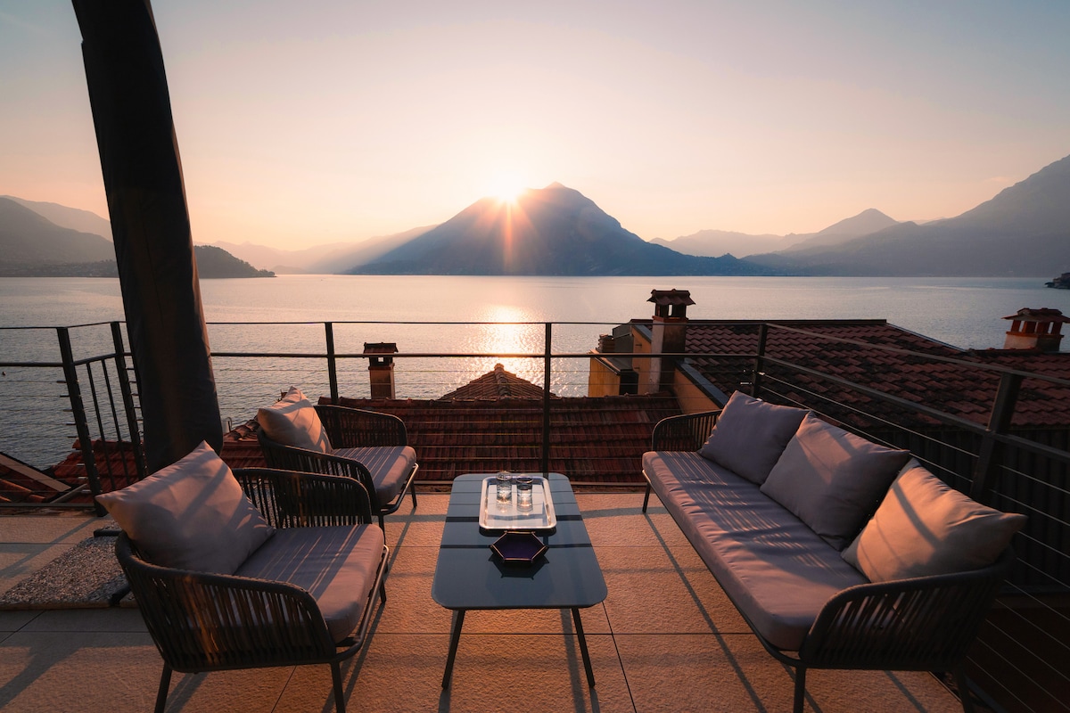 An outdoor seating area is depicted on a terrace overlooking the tranquil lake and surrounding mountains. Two cushioned lounge chairs are positioned around a small glass table, capturing the serene colors of a sunset reflecting on the water.