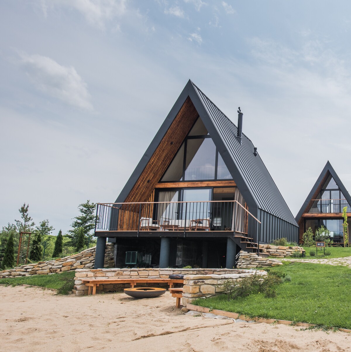 A modern triangular cabin features large glass windows, allowing for ample natural light. A spacious balcony with outdoor seating overlooks the sandy area, while a fire pit is placed in front of the cabin. Lush greenery surrounds the structure, enhancing its connection to nature.
