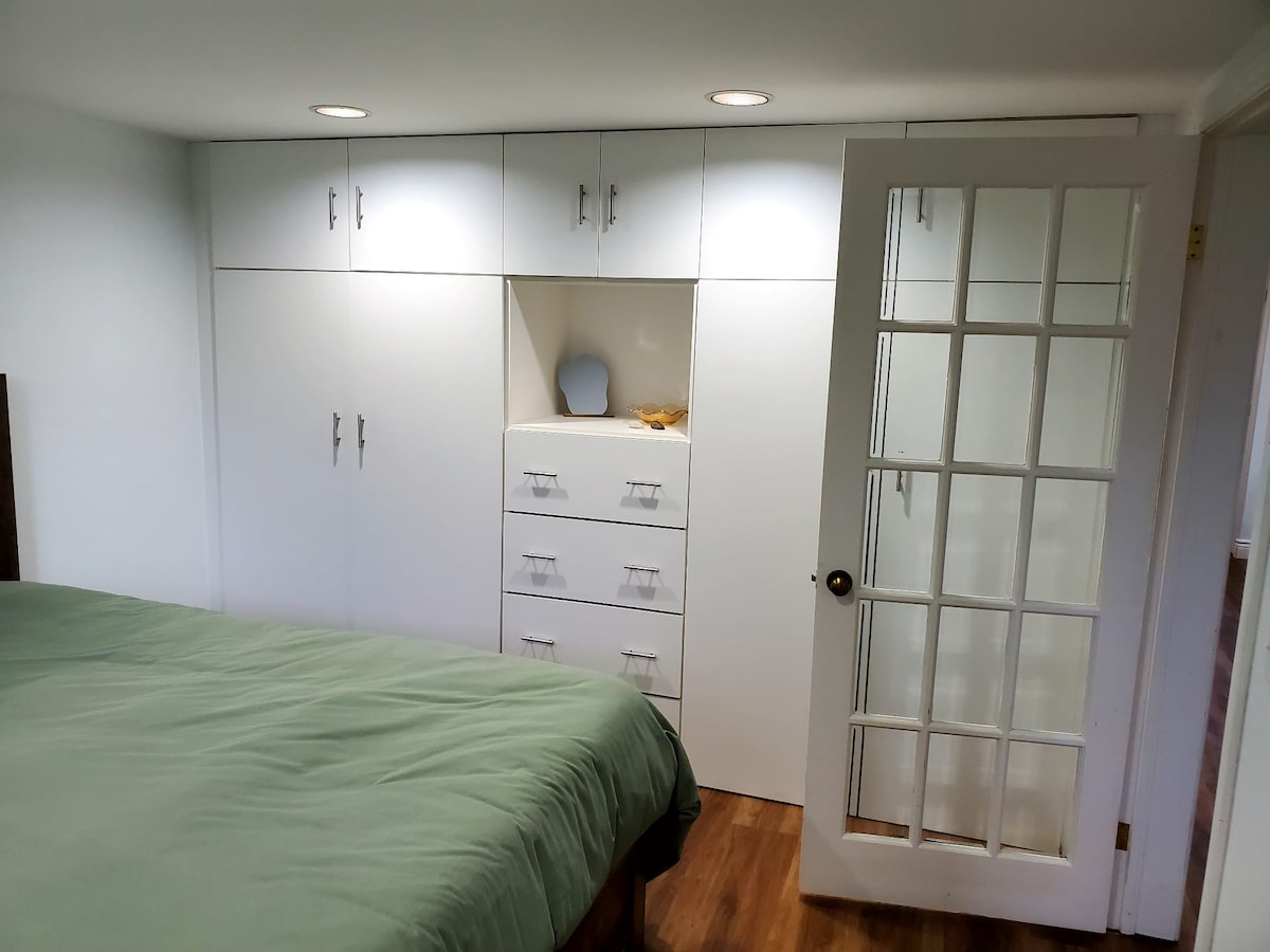 A cozy sleeping area features a spacious bed with a green blanket. Opposite the bed, sleek built-in cabinetry and a set of drawers are visible, providing ample storage. A glass-paned French door leads to another space, allowing natural light to enhance the room.