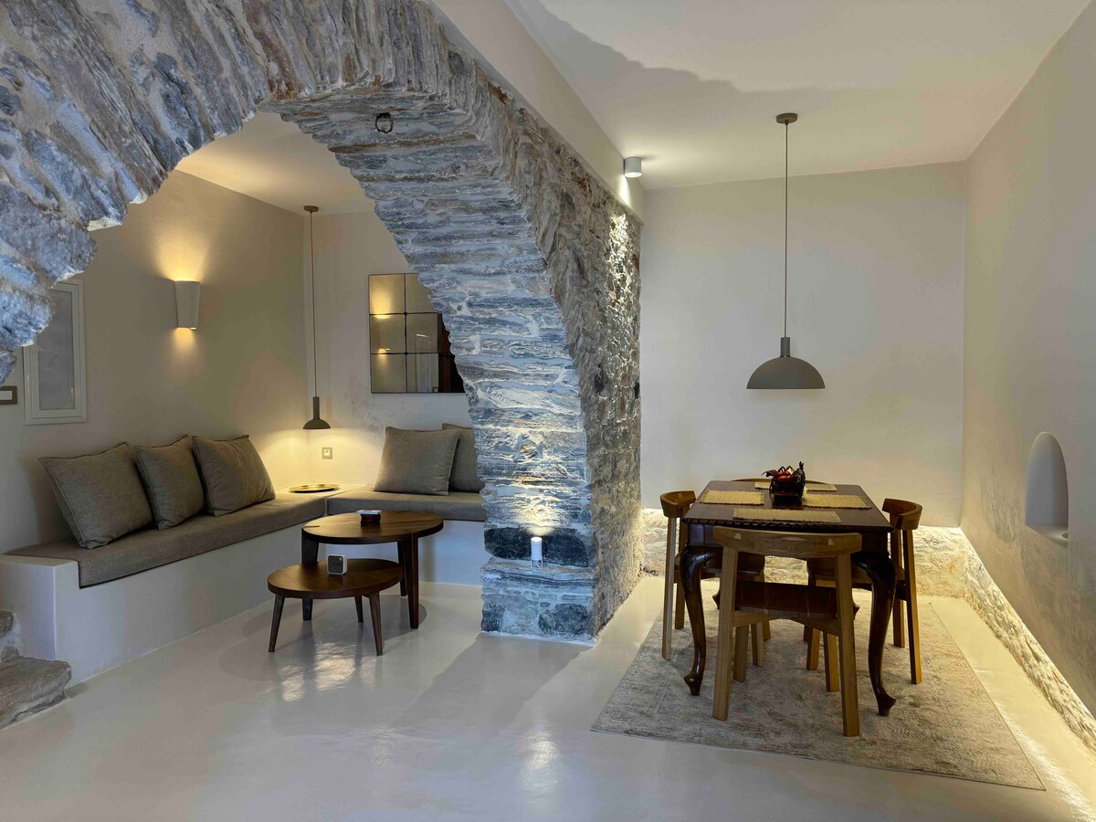 A stone archway leads into an open living and dining area. Soft seating is arranged beside a textured wall, while a wooden dining table is surrounded by matching chairs. Soft lighting illuminates the space, enhancing the clean and modern lines of the interiors.