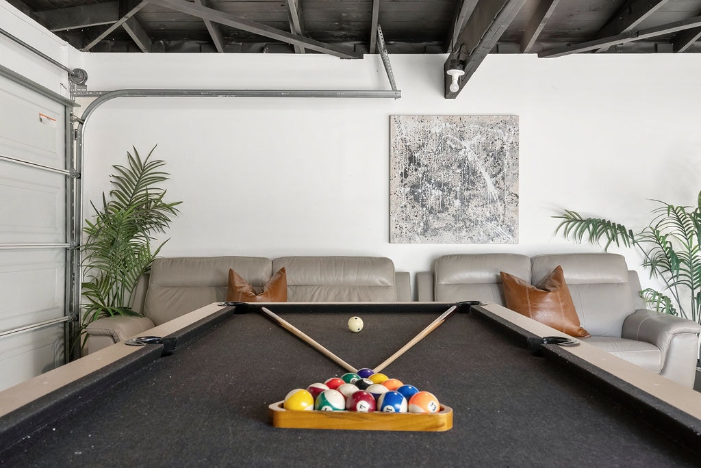 A billiards table is centrally positioned in the game room, with a rack of colorful pool balls set for play. Light-colored sofas are arranged nearby, complemented by decorative cushions and green plants adding a touch of nature to the space.