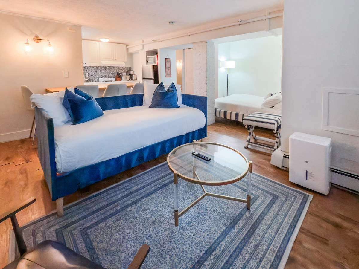 A shared living space features a cozy daybed adorned with blue cushions. A round glass coffee table rests on a patterned rug, adding comfort. In the background, a light-filled bedroom is visible, showcasing a striped bedspread and additional seating.