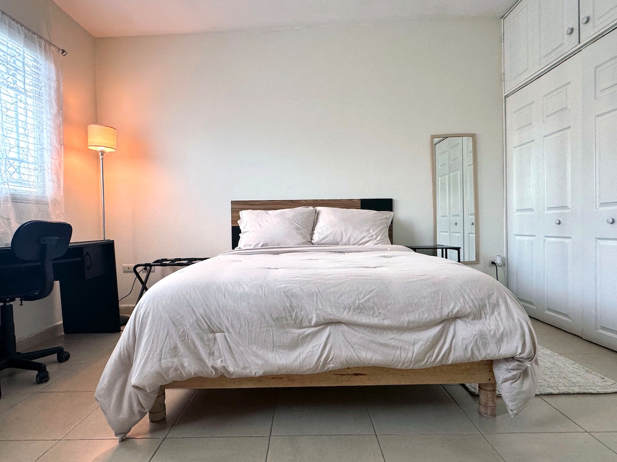 A spacious bedroom features a large bed with light bedding. A desk and chair are positioned nearby, while a lamp provides soft lighting. Storage is available in a cabinet with multiple doors, and natural light filters through a window with sheer curtains.