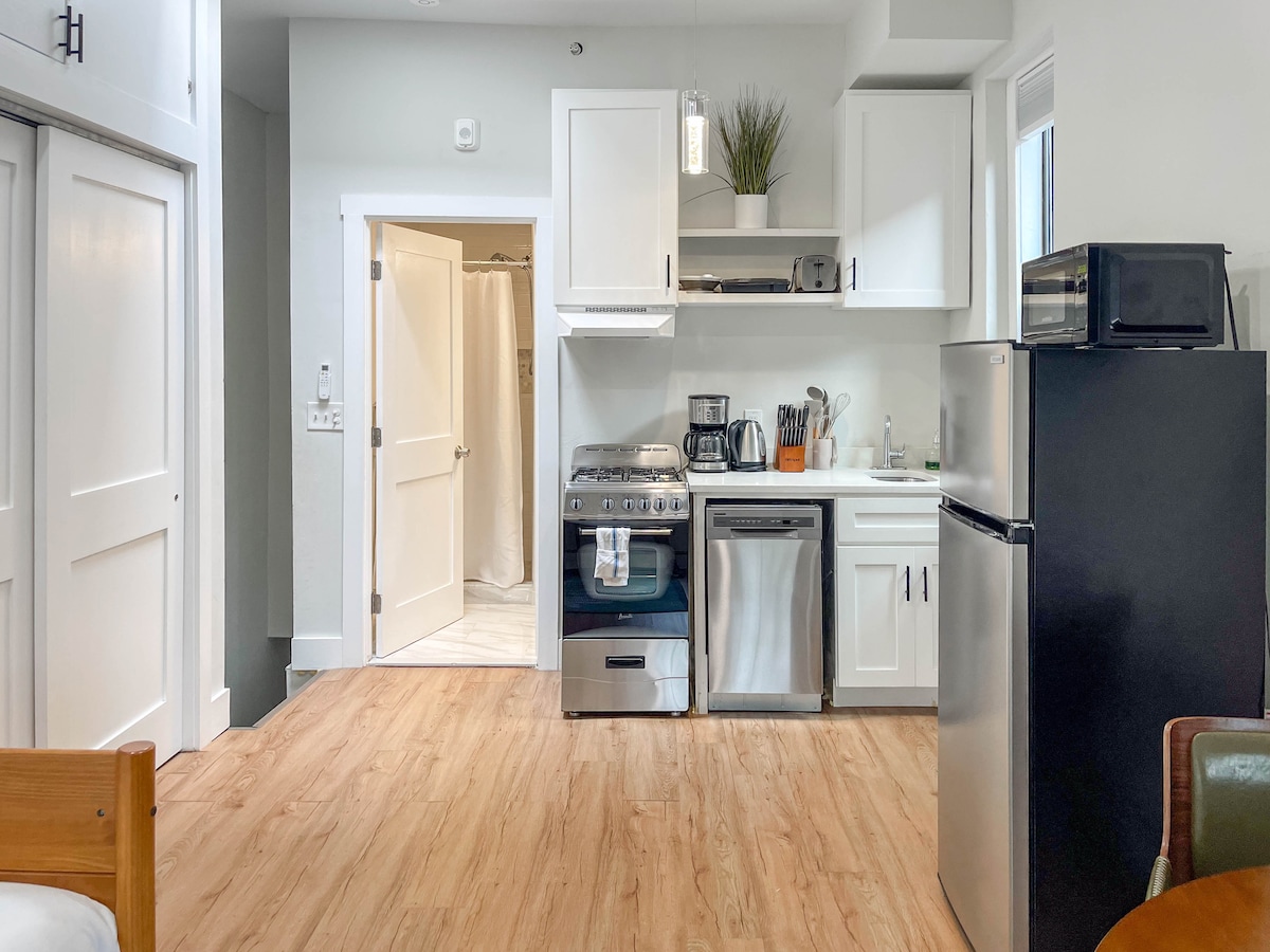 A compact kitchen area is equipped with modern appliances, including a stove, microwave, and refrigerator. Light wood flooring provides a warm feel, while white cabinetry complements the space. A door leads to a bathroom, and a glimpse of a cozy sleeping area is visible in the foreground.