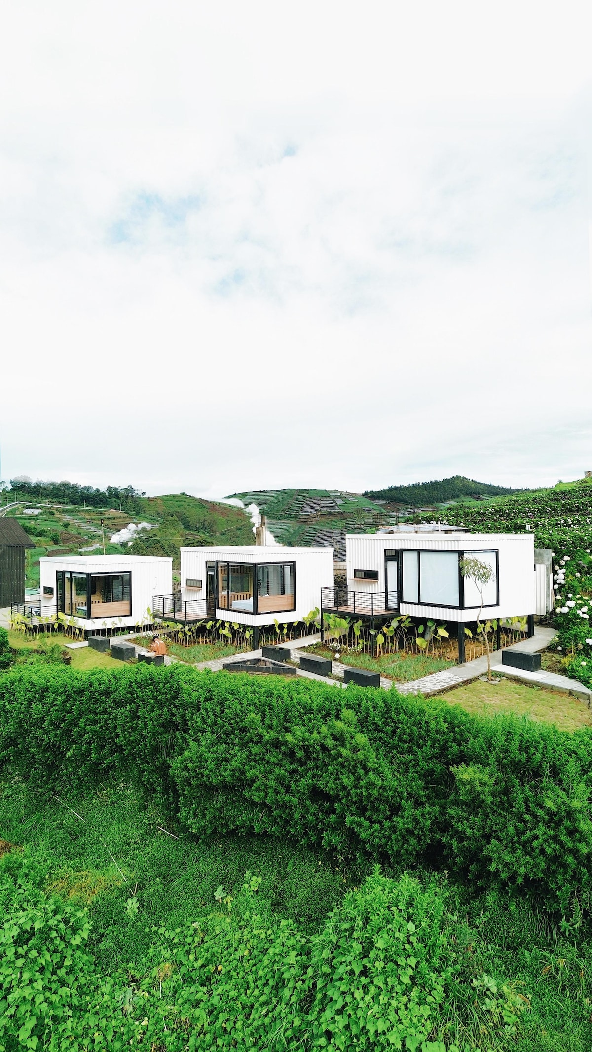 Three modern accommodations are nestled amidst lush greenery, framed by gentle hills in the background. Each unit features large windows, allowing for ample natural light while providing views of the surrounding landscape. Outdoor seating areas are visible in front, enhancing the connection to nature.