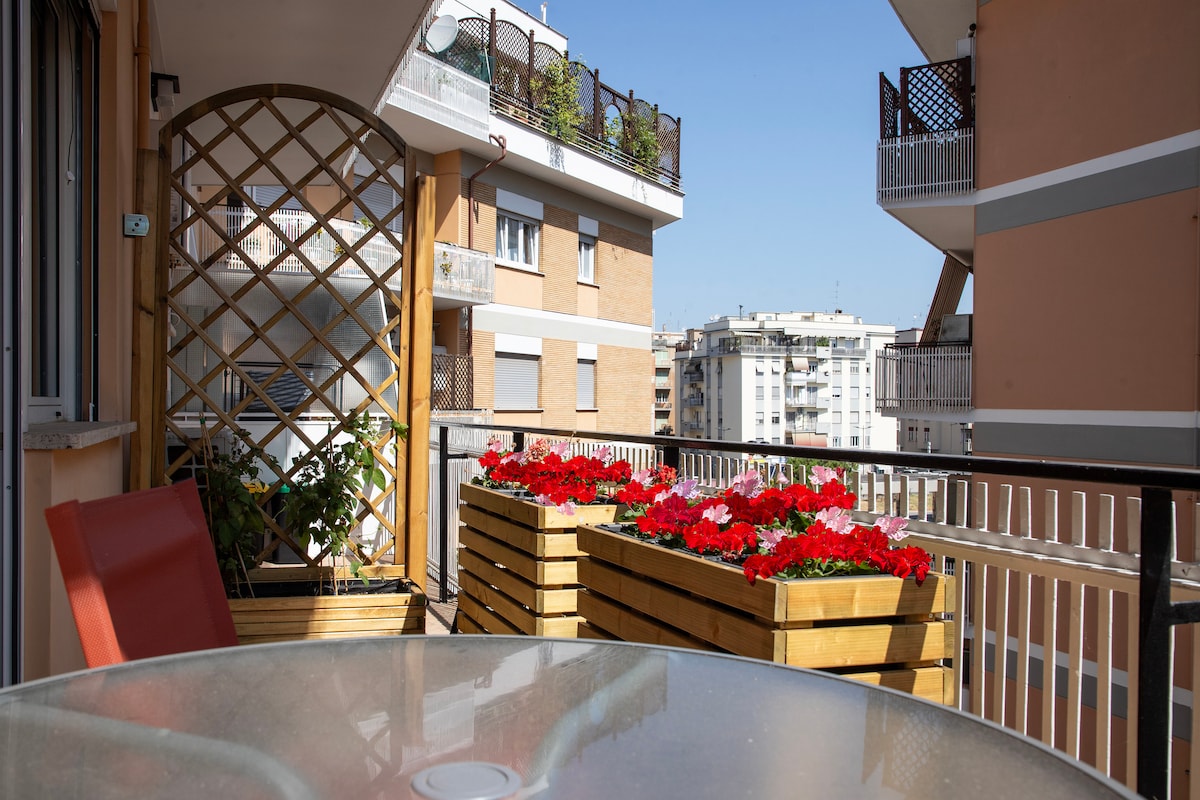 A terrace area is seen with wooden planters filled with vibrant red flowers. A round glass table is positioned on the terrace alongside a red chair. The backdrop features nearby buildings under a clear blue sky.