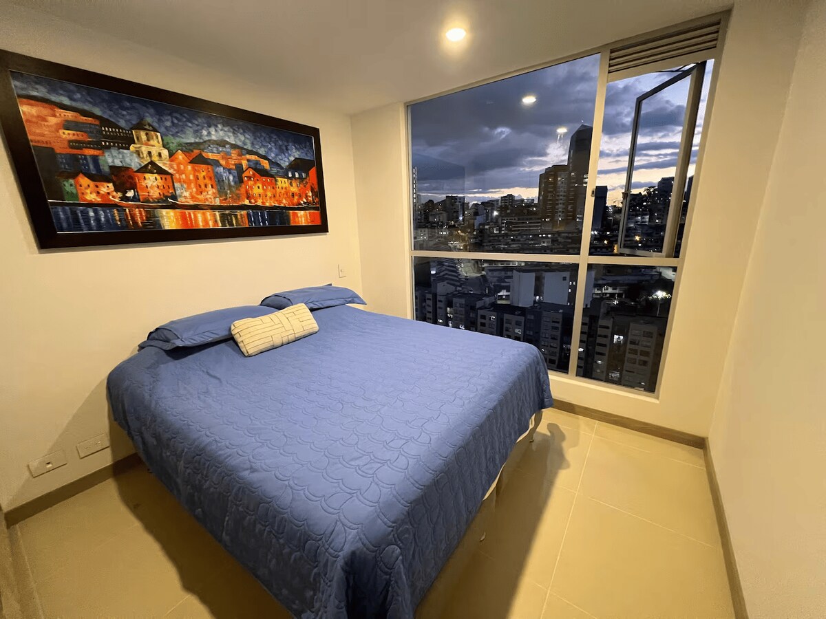 A comfortable bedroom features a spacious bed covered with a blue quilt. A vibrant painting adorns the wall above the bed, while large windows offer a view of the city skyline at dusk, with clouds creating a dramatic backdrop.