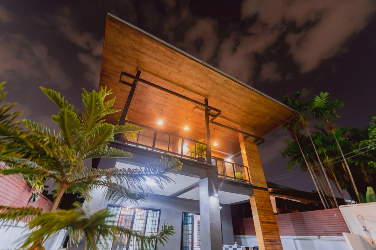 A modern two-story house features a wooden overhang and large windows that invite natural light. Soft lighting enhances the architectural details, while surrounding palms create a relaxed atmosphere. The exterior is designed with a contemporary style, emphasizing both comfort and elegance.