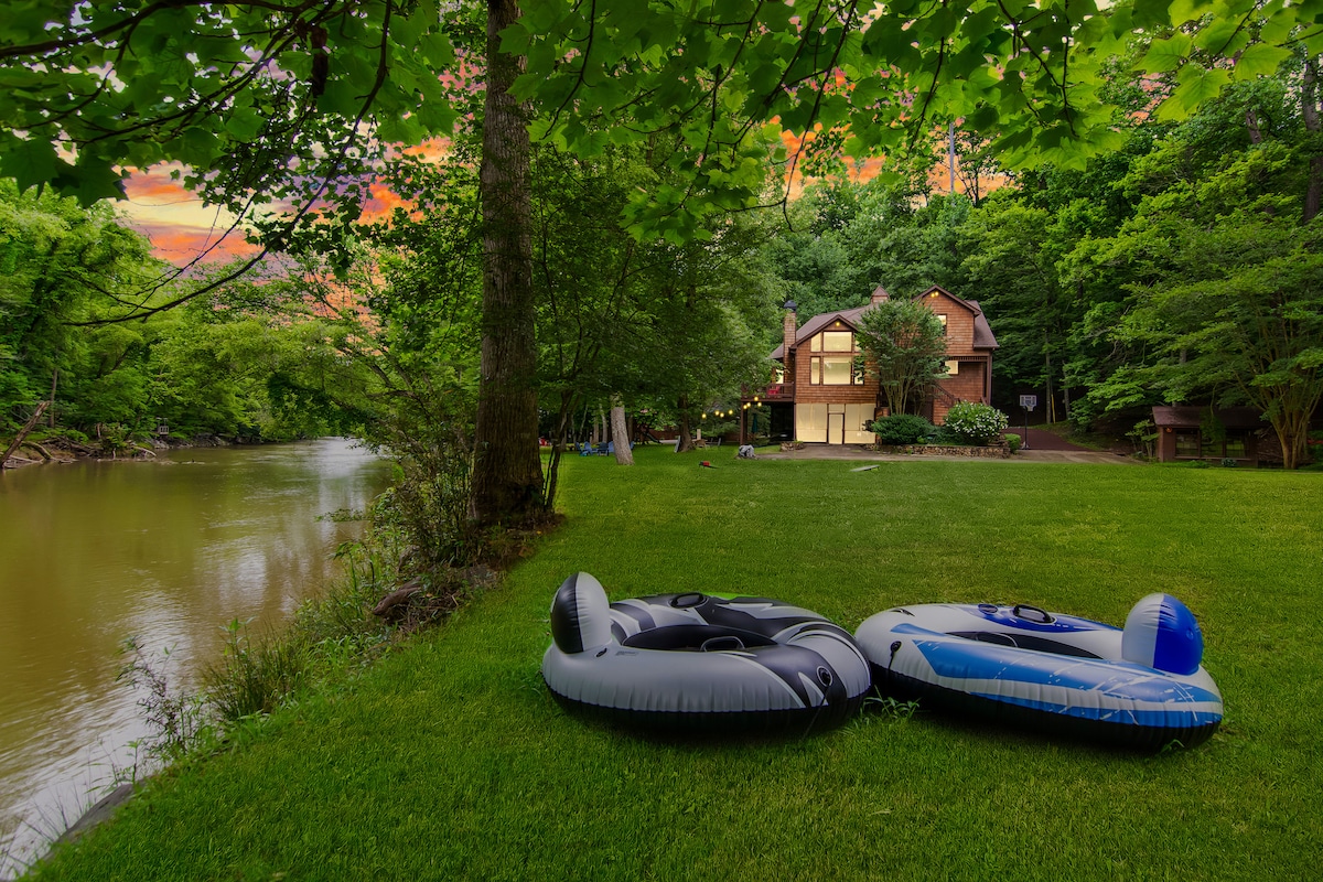 Two inflatable tubes rest on a grassy area beside a calm river. The cabin, featuring large windows, is visible in the background, surrounded by lush trees. A serene sunset adds warm colors to the sky, enhancing the natural setting.