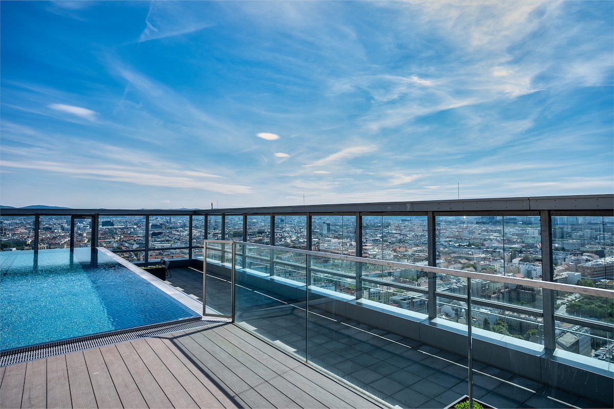A rooftop area showcases a modern pool overlooking a panoramic city view. Glass panels surround the space, providing safety while allowing for unobstructed sightlines. Wooden decking complements the sleek design, creating an open and airy environment.