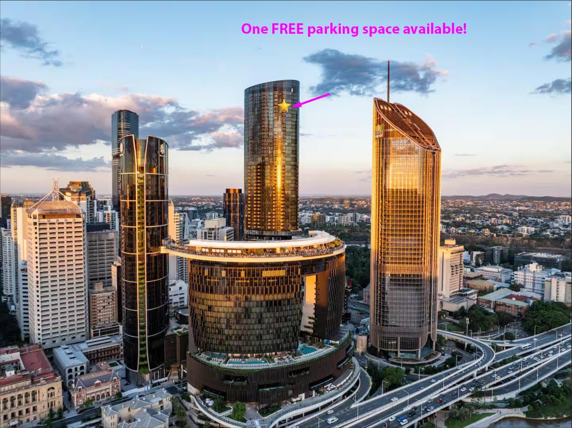 An aerial view captures the modern skyline of Queen’s Wharf, featuring gleaming high-rise buildings bathed in warm sunlight. The surrounding area showcases a network of roads and green spaces, emphasizing the vibrant urban environment. One highlighted parking space is noted as available.