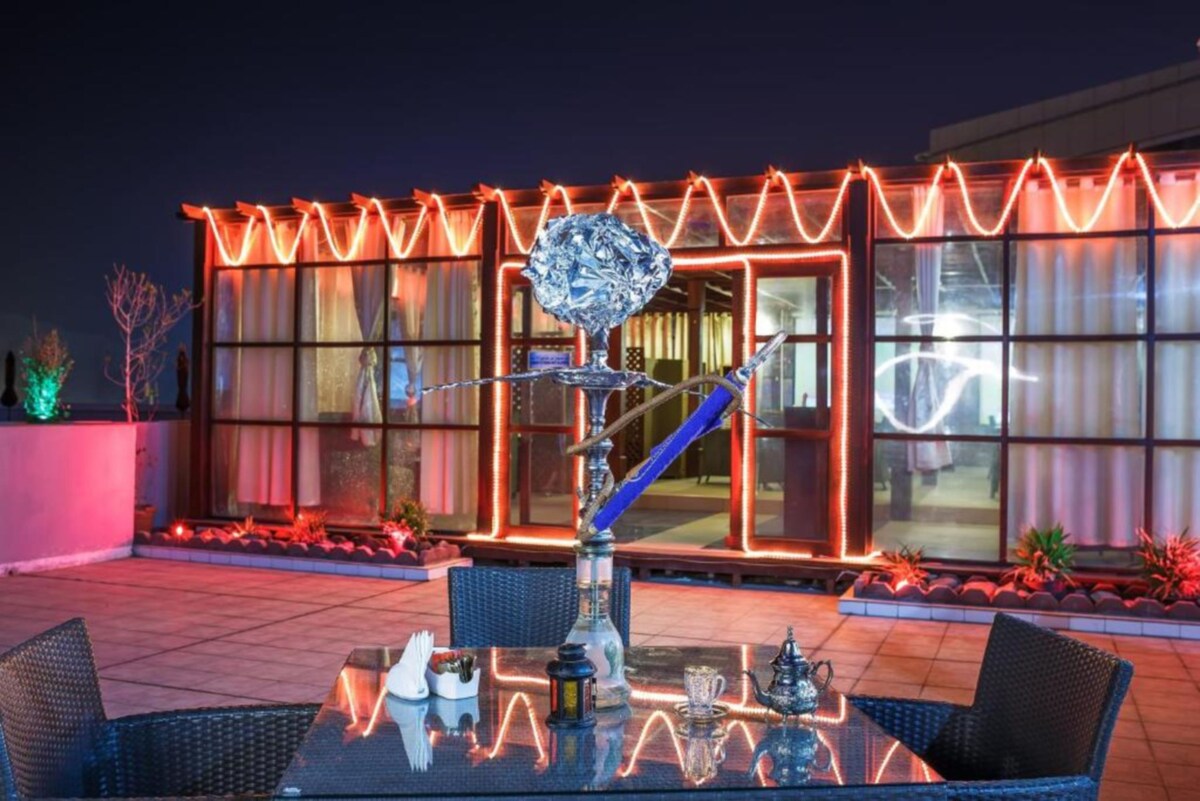 A rooftop terrace is presented with a glass table and dark wicker chairs, featuring a decorative water pipe and small cups. Colorful lights outline the adjacent glass structure, enhancing the evening atmosphere. Potted plants are visible along the patio, adding greenery to the space.