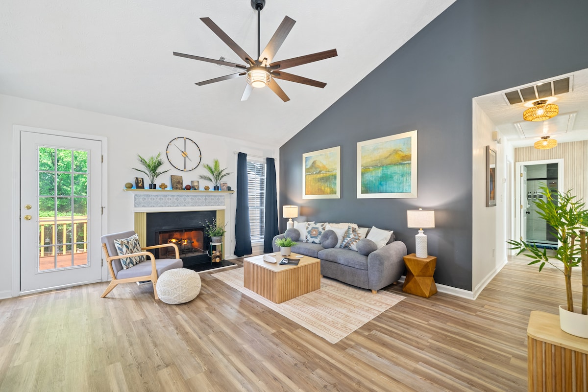 A living area features a gray couch complemented by decorative pillows, a light wood coffee table, and a round floor pouf. Artwork adorns the walls, with a modern light fixture above. A fireplace is framed by a white mantel, and large windows provide views of the outdoor greenery.