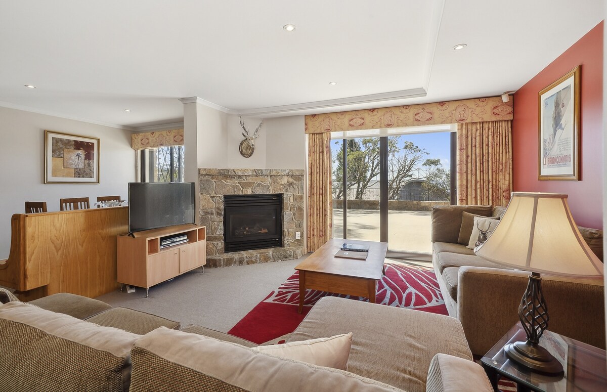A spacious living area is presented with a large brown sofa, soft lighting, and a cozy fireplace surrounded by stone. Large glass doors lead to a private outdoor space, while a dining area is visible in the background, accented by warm wooden tones.