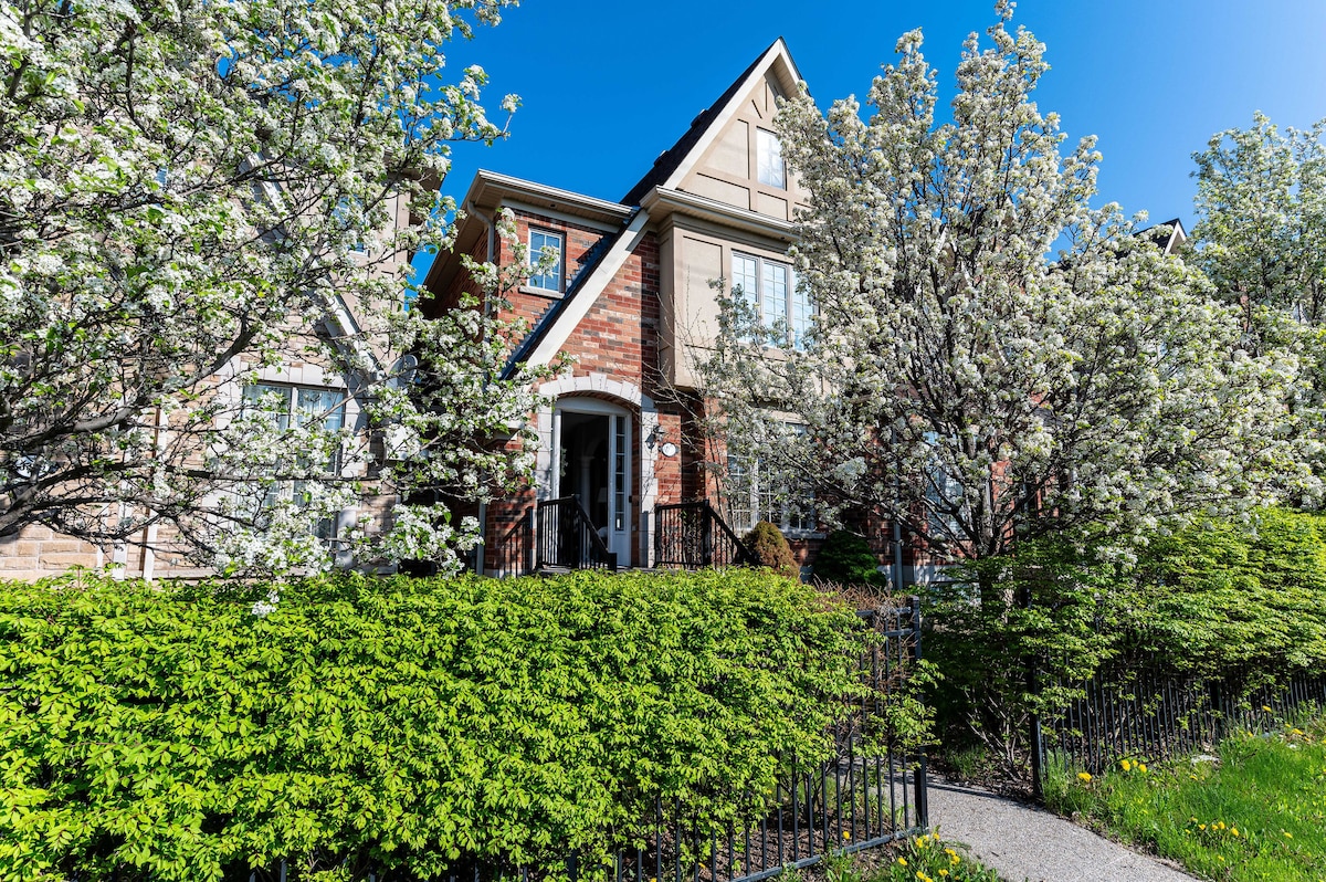 The townhouse's exterior is framed by blooming trees and manicured shrubs. A pathway leads to the inviting entrance, characterized by a central door flanked by large windows. The architectural design features a cozy gabled roof and a blend of brick and siding materials.