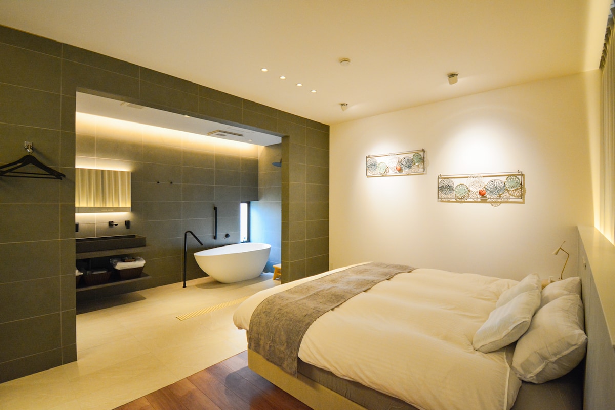 A modern bedroom includes a spacious bed with soft linens. A minimalist design features soothing neutral tones on the walls, with softly illuminated art pieces. A large window provides natural light, and a bathroom area is visible, highlighted by a freestanding tub and contemporary fixtures.