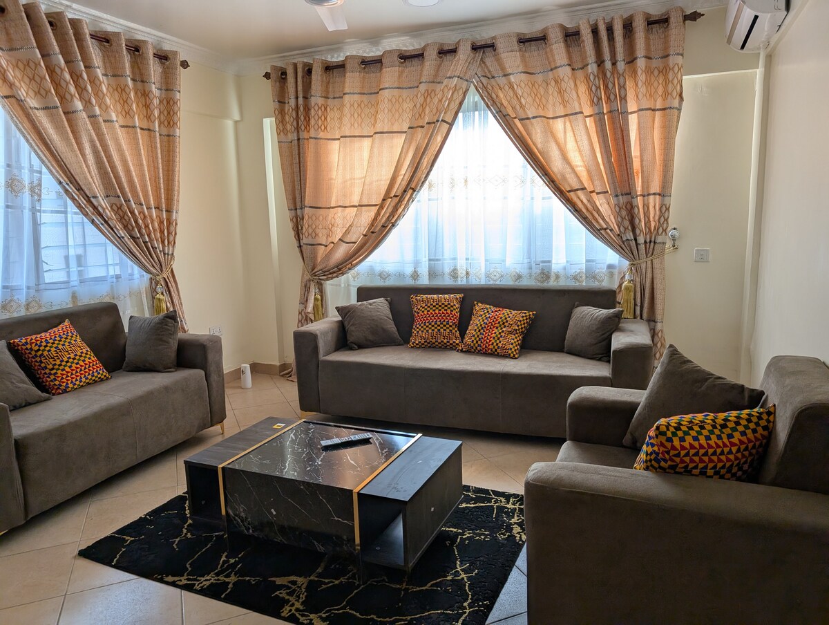 A comfortable seating area features two large sofas and one armchair, all arranged around a central coffee table. Decorative cushions add color to the neutral-toned upholstery. Light filters through sheer curtains, illuminating the space, with patterned drapes framing the windows.