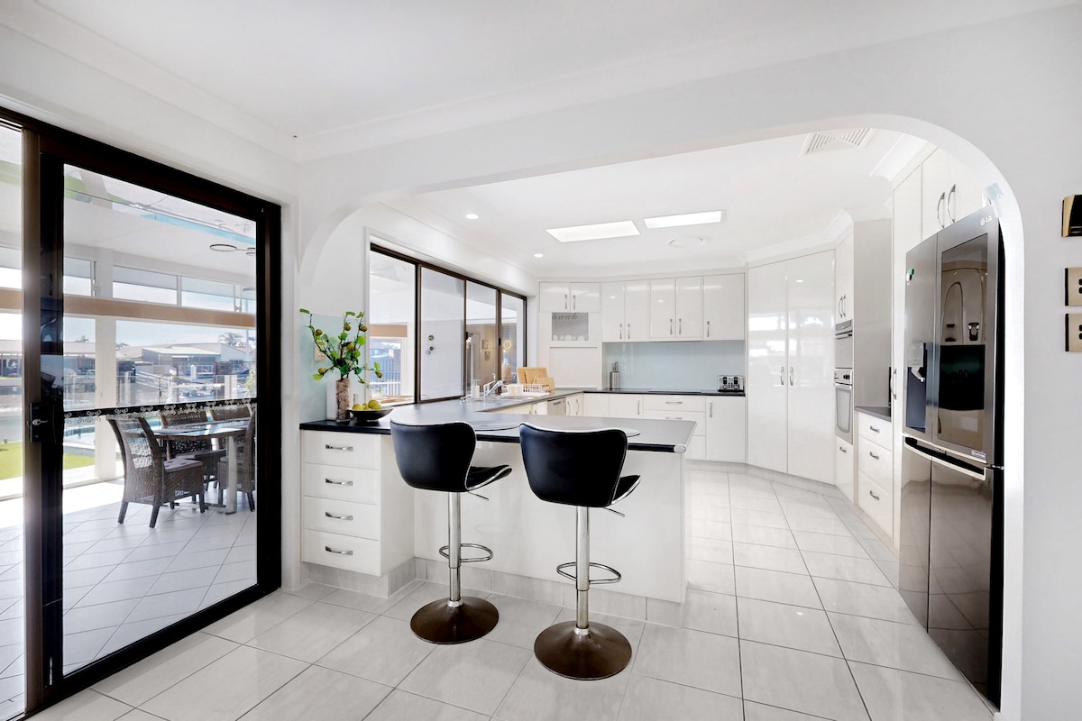 A modern kitchen features sleek white cabinetry and a spacious island with two black bar stools. Large windows provide ample natural light, creating an open feel. Stainless steel appliances and a simple vase of greenery enhance the contemporary design.
