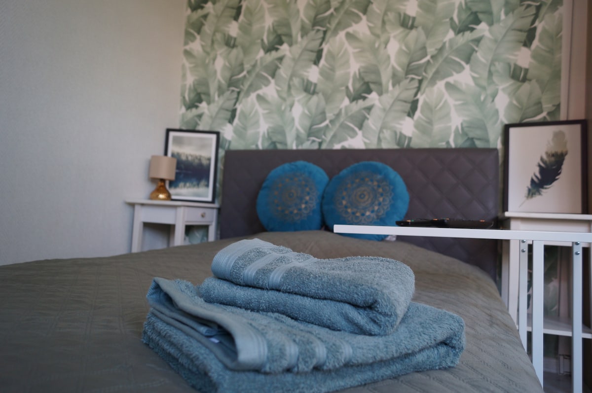 A cozy bed is adorned with plush pillows and a calming blanket. On the bedside table, a lamp provides soft light. Fresh towels are neatly arranged at the foot of the bed. Botanical wallpaper enhances the room's tranquil atmosphere.