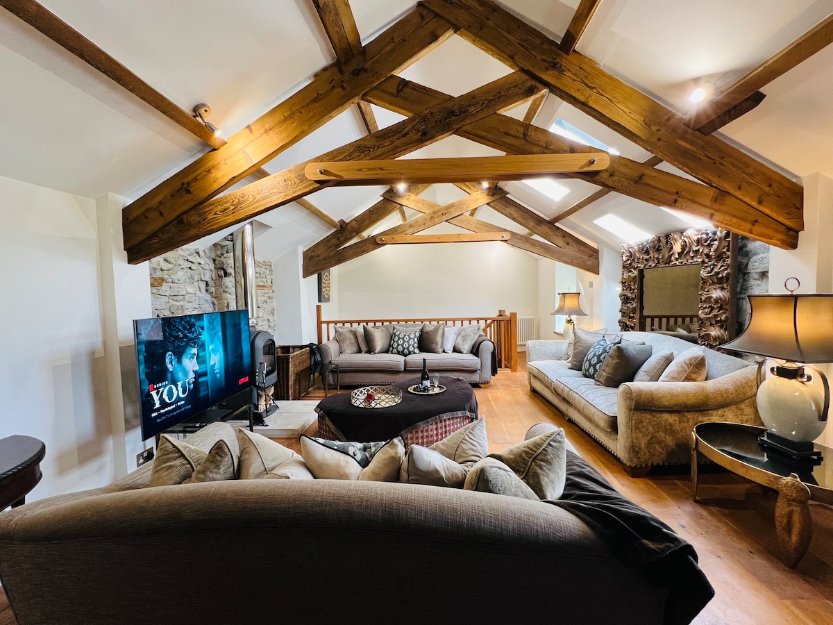 The spacious lounge offers a warm and inviting space, featuring exposed wooden beams and large windows that bring in natural light. Comfortable seating arrangements include a sofa and a cozy armchair, complemented by a stylish coffee table. A smart TV is positioned for entertainment.