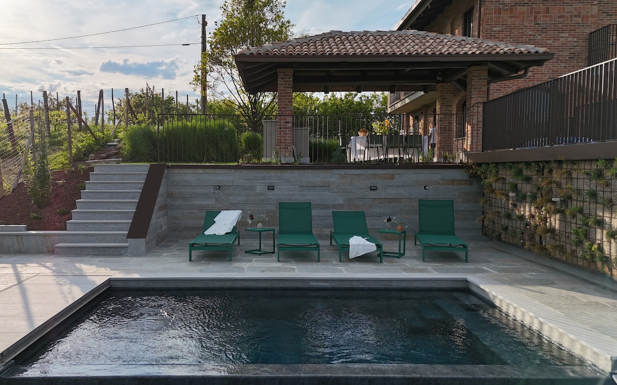 A serene pool area features a rectangular swimming pool surrounded by four green lounge chairs. A covered outdoor kitchen is visible in the background, along with well-maintained gardens and distant vines, offering a peaceful space for relaxation.