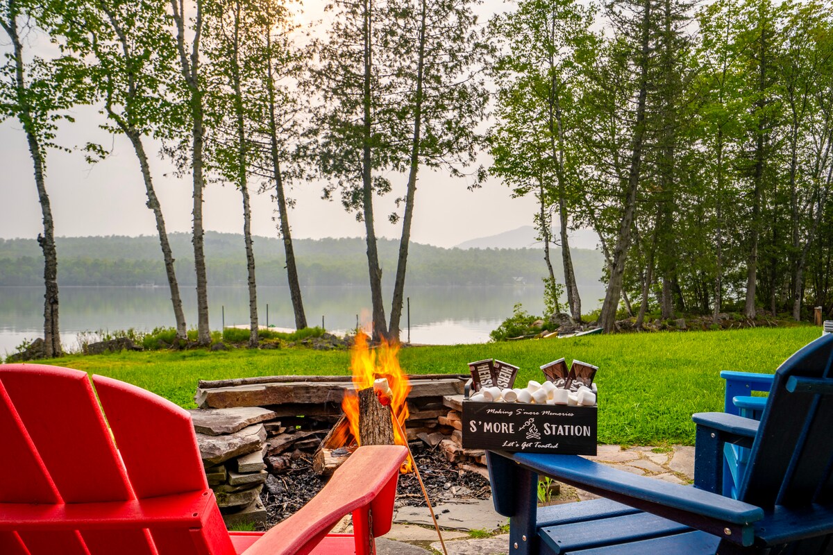 A cozy fire pit is surrounded by colorful adirondack chairs, inviting relaxation near the water's edge. A tranquil view of Shin Pond and trees is visible, creating a serene atmosphere for gatherings. A sign for a s'mores station complements the outdoor setting.