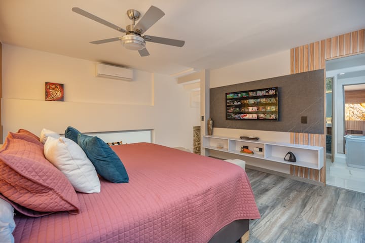 חדר שינה ראשי נעים עם מיטת קינג סייז, מאוורר תקרה וטלוויזיה חכמה מותקנת על הקיר למנוחה אולטימטיבית.