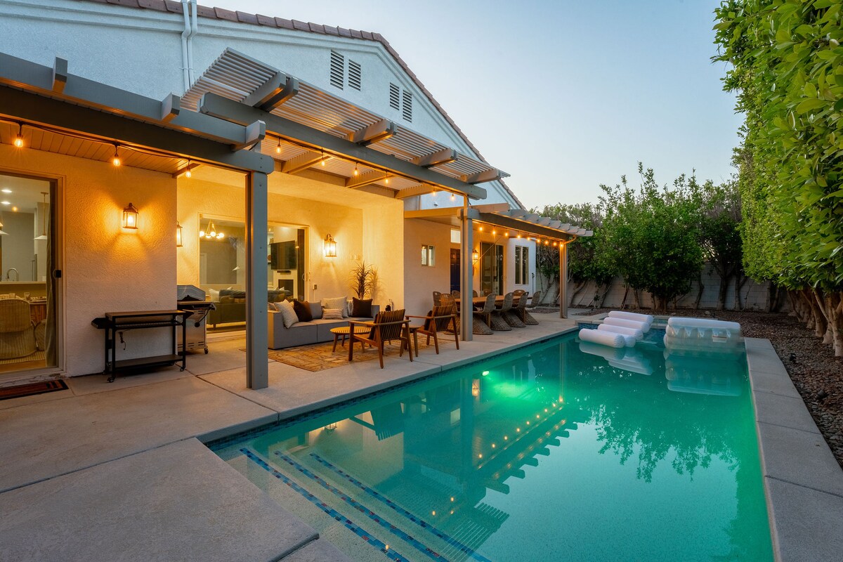 An outdoor area features a private swimming pool with clear water, reflecting the surrounding space. Comfortable seating arrangements and a dining area are shaded under a pergola, adorned with soft lighting. Lush greenery lines the perimeter, adding a sense of privacy and tranquility.