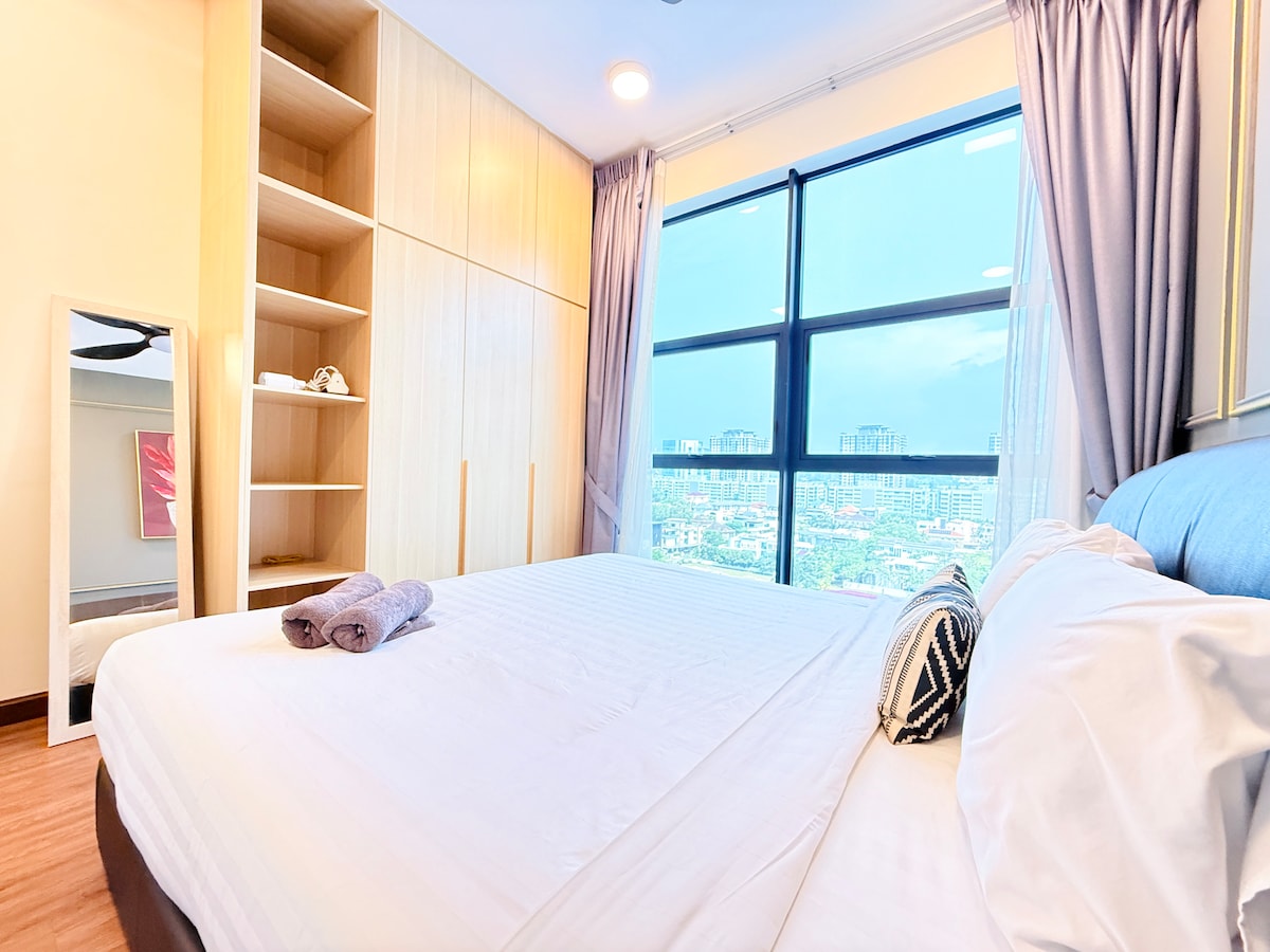 The master bedroom features a spacious bed with hotel-quality bedding and decorative pillows. Floor-to-ceiling windows provide views of the surrounding area, with sheer curtains allowing natural light. A built-in wardrobe offers storage options, while soft neutral tones create a calming atmosphere.