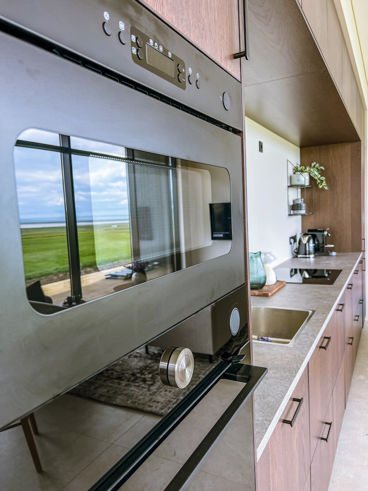 The image captures a sleek kitchen featuring modern appliances. A built-in oven is prominently displayed with a glass door, and a functional sink is situated beside it. Dark cabinetry complements the neutral countertops, while large windows provide views of the surrounding landscape.