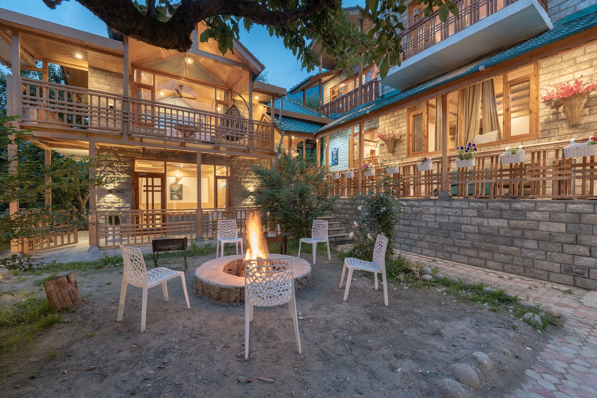 A charming outdoor area is featured, hosting a circular stone fire pit surrounded by four white chairs. The villa's exterior showcases wooden balconies and stone accents, illuminated by soft evening light, creating a cozy gathering space amidst the lush green surroundings.