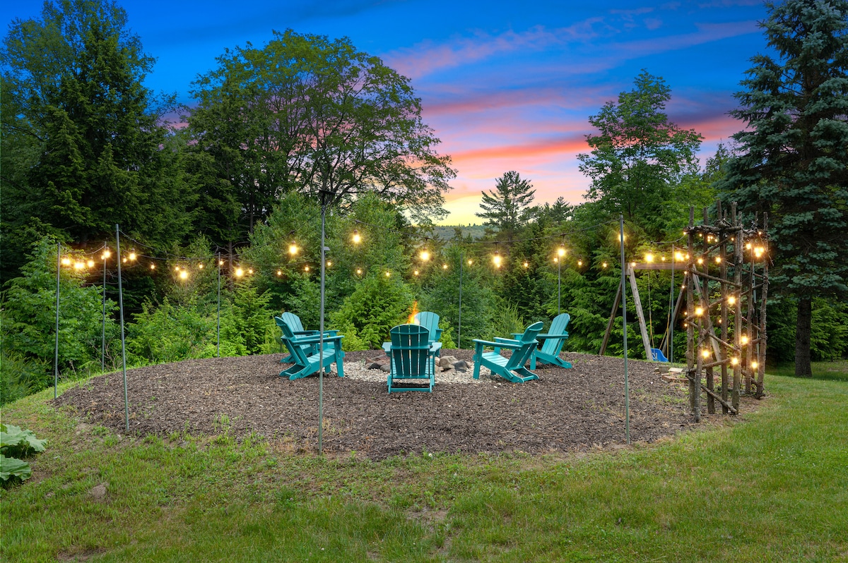 A circular fire pit area is surrounded by vibrant greenery and features Adirondack chairs arranged around the fire. Outdoor string lights create a warm glow above, illuminating the space as dusk approaches, inviting gathering and conversation under the sky.