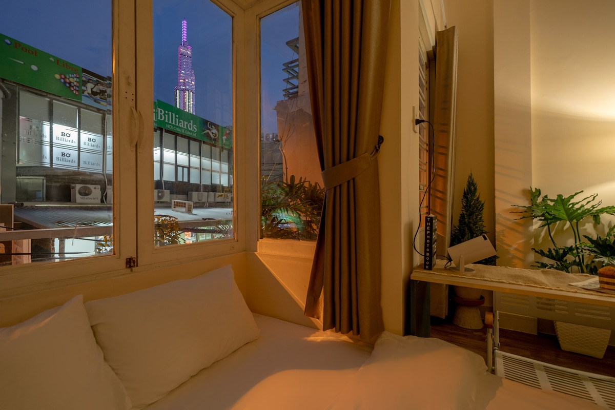 A cozy corner of the room features a spacious bed with soft white linens. Natural light filters through the window, revealing a vibrant city view highlighted by the glowing Landmark 81. A small workspace and indoor plants enhance the serene atmosphere.