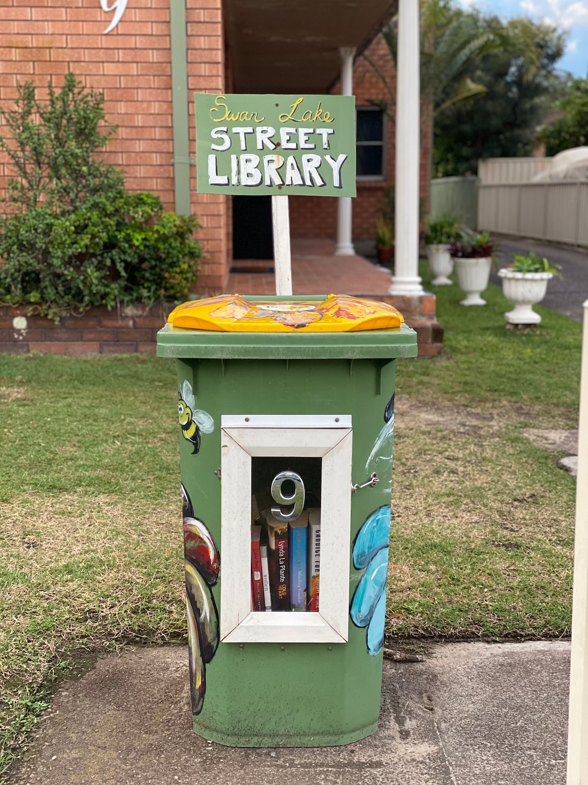 A colorful street library is showcased, featuring a green painted structure with artistic designs. A sign labeled 'Swan Lake Street Library' is perched on top. Books are visible through a window at the front, inviting community members to borrow and exchange reading materials.