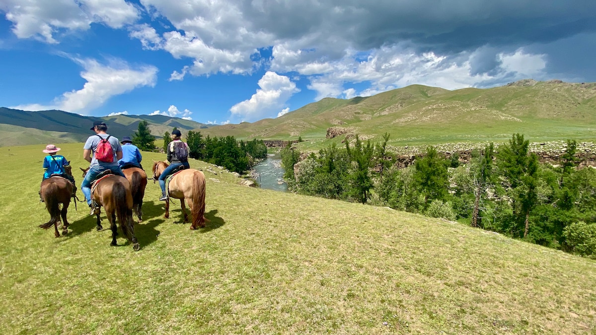 A group of four individuals on horseback is seen riding along a grassy landscape. In the background, a river flows beside green trees against rolling hills under a partly cloudy sky.