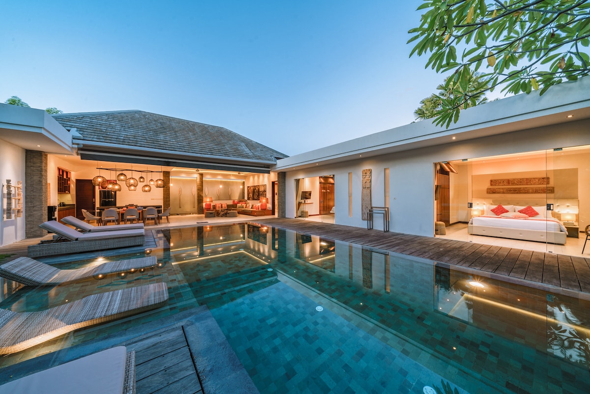 An open-plan villa is showcased, with a large private pool reflecting the evening sky. Sliding glass doors create an inviting connection between indoor living areas and the outdoor space. Comfortable lounge chairs are arranged along the poolside, highlighting the serene atmosphere.