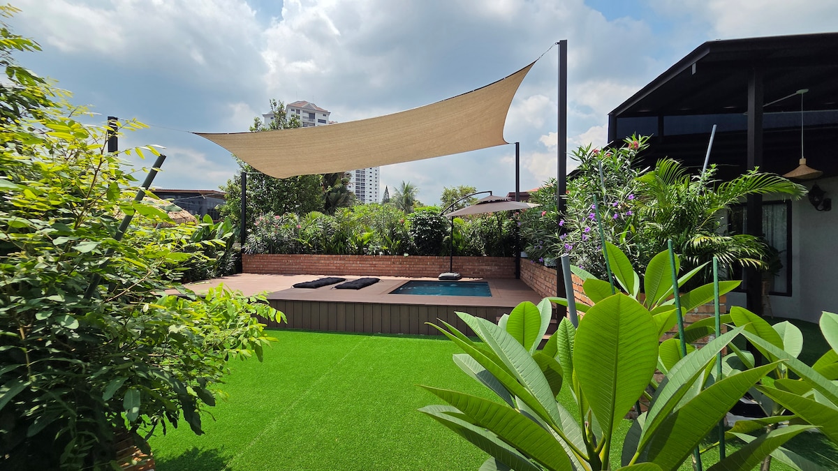 A serene outdoor area is surrounded by lush greenery, featuring a sunshade that provides relief from sunlight. A small pool is visible alongside lounge chairs, inviting relaxation. The well-kept grass adds to the tranquil atmosphere of this lush retreat.
