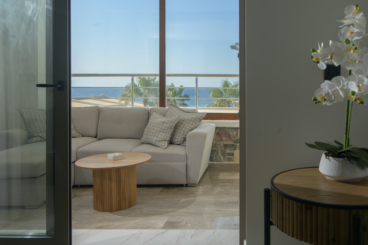 A light-filled seating area features a modern gray sofa adorned with patterned cushions, paired with a small round wooden table. Large windows reveal a stunning sea view, enhancing the space's open feel while allowing natural light to flood in.