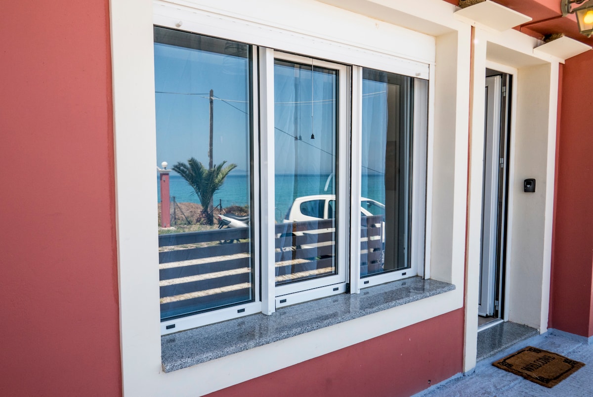 A row of three large windows with white frames offers a clear view of the sea. The light reflects off the glass, revealing a glimpse of the vibrant blue water and palm trees outside, set against the red exterior of the building.