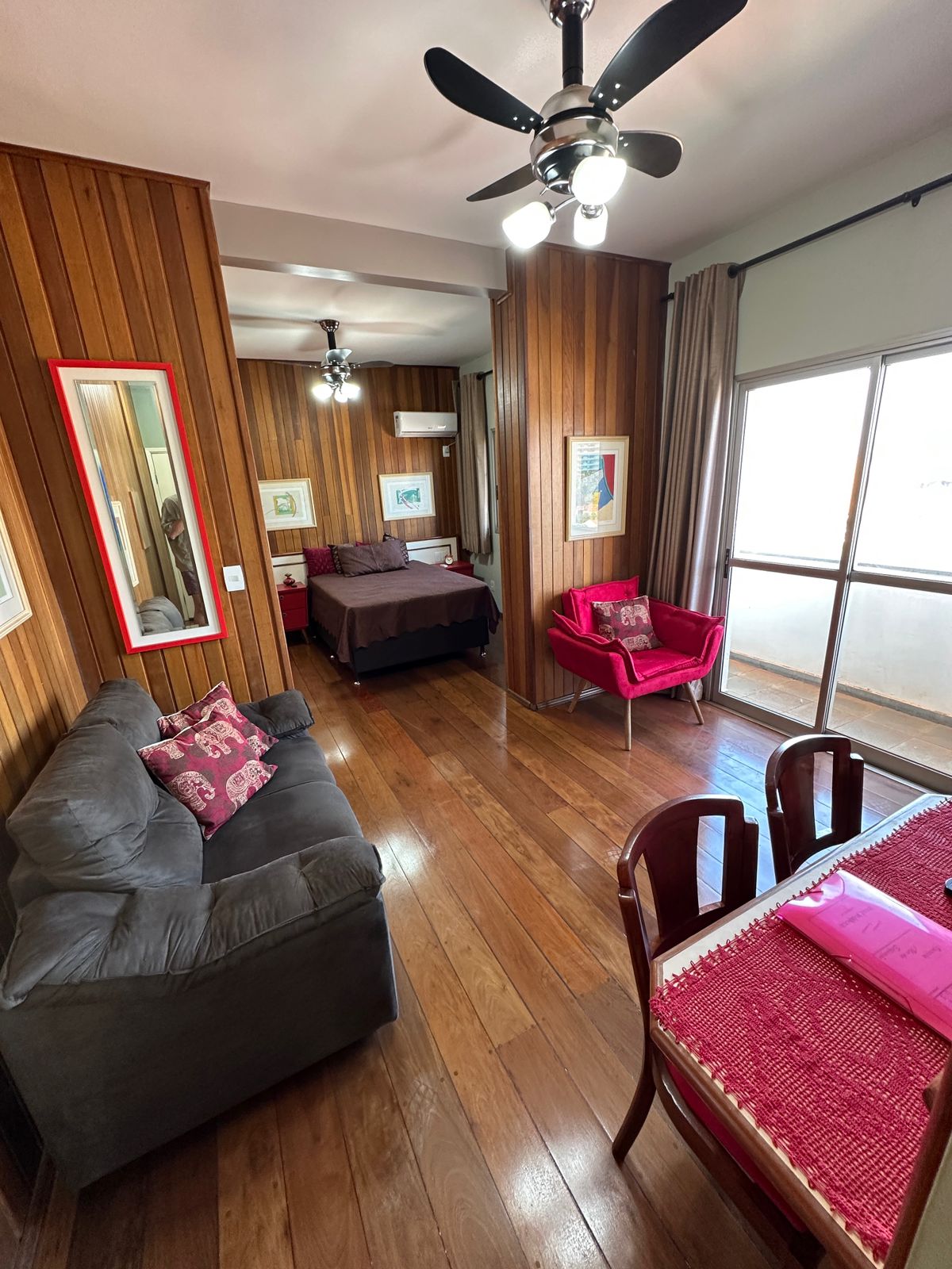 A cozy interior is showcased with wooden walls and flooring. A comfortable gray sofa and two vibrant red chairs are positioned around a dining table. The spacious bed is visible in the background, complemented by natural light streaming through large windows.